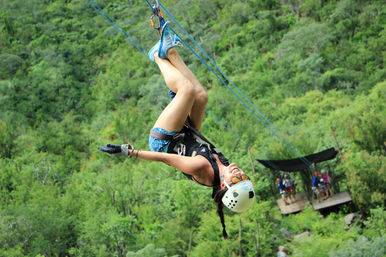 Grinning zipline rider in helmet and harness hangs upside down on a blue cable over a lush green forest canopy — outdoor canopy tour with wooden launch platform and people below