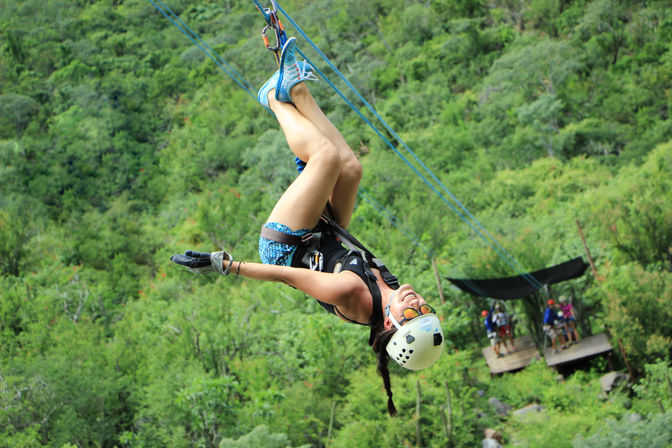 Grinning zipline rider in helmet and harness hangs upside down on a blue cable over a lush green forest canopy — outdoor canopy tour with wooden launch platform and people below