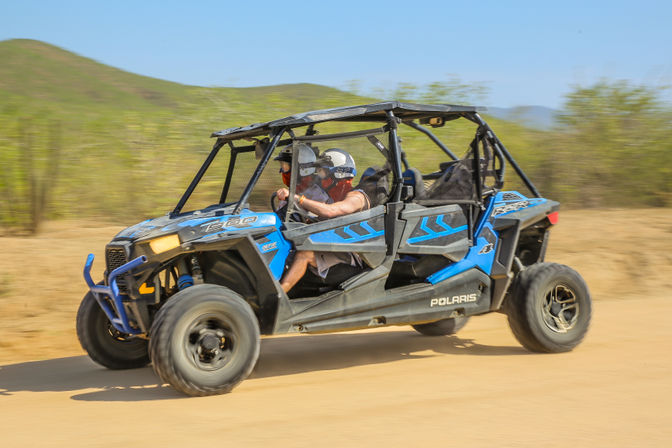 Blue four-seat off-road UTV (side-by-side) with two helmeted riders speeding along a dusty desert trail, motion-blurred background