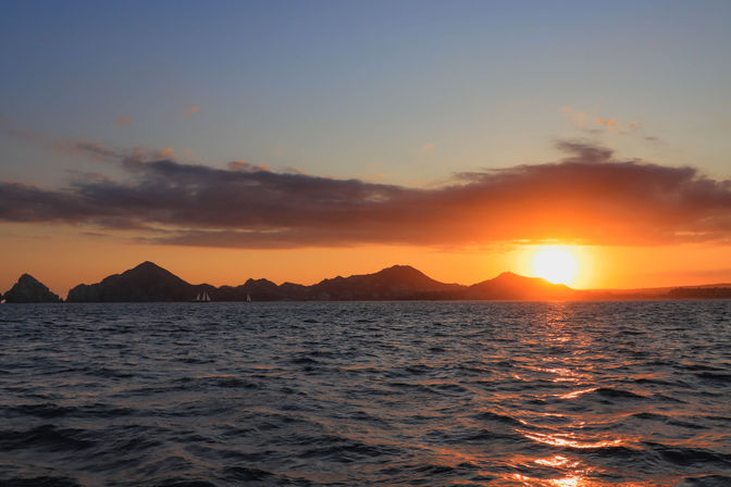 Sunset over open sea with the orange sun dipping behind silhouetted coastal hills, rippled water reflecting golden light and a few sailboats on the horizon