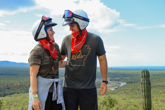 Two helmeted hikers wearing red bandanas smile on a sunny desert overlook by a tall cactus, with winding river, green scrubland and distant ocean horizon