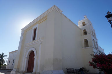 Sunlit white colonial-style church with arched wooden door and multi-level bell tower against a clear blue sky, benches and a flowering shrub in the foreground