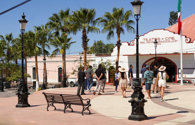 Sunny Mexican town square with palm trees and vintage lampposts, tourists in summer clothes and sun hats strolling past a white-and-red historic theater facade, ornate bench in the foreground and a Mexican flag overhead.