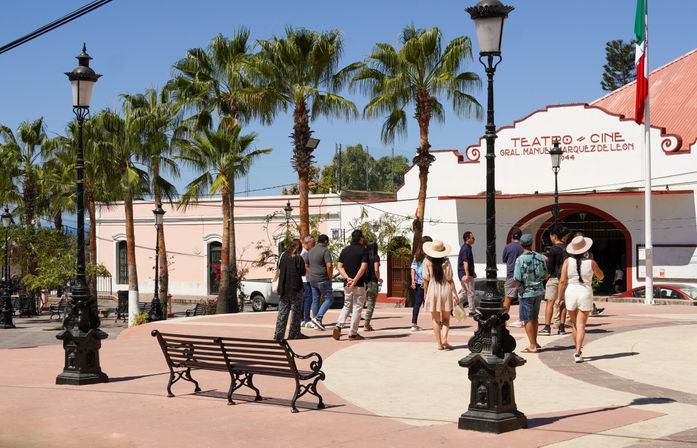 Sunny Mexican town square with palm trees and vintage lampposts, tourists in summer clothes and sun hats strolling past a white-and-red historic theater facade, ornate bench in the foreground and a Mexican flag overhead.