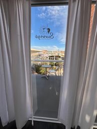 View through sheer white curtains to a small balcony with a round table and metal chair, sun-themed logo decal on the glass and a sunny urban plaza with blue sky beyond.