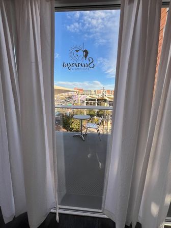 View through sheer white curtains to a small balcony with a round table and metal chair, sun-themed logo decal on the glass and a sunny urban plaza with blue sky beyond.