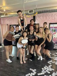 Group of friends posing at a pole fitness studio with pink faux-fur walls, holding bachelorette props and surrounded by scattered play money — upbeat pole party photo.