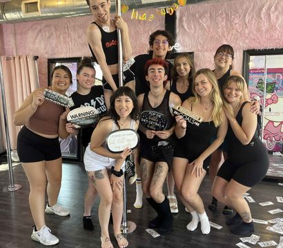 Group of friends posing at a pole fitness studio with pink faux-fur walls, holding bachelorette props and surrounded by scattered play money — upbeat pole party photo.