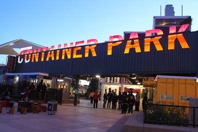 Entrance to an urban outdoor marketplace made from repurposed shipping containers with large glowing orange letters on the container facade at dusk, clusters of people gathered below.