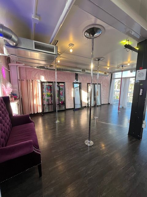 Bright pole fitness studio interior with multiple chrome dance poles, full-length mirrors, dark wood flooring, tufted purple velvet sofa, pink textured walls, exposed ductwork, and floor-to-ceiling windows with natural light.