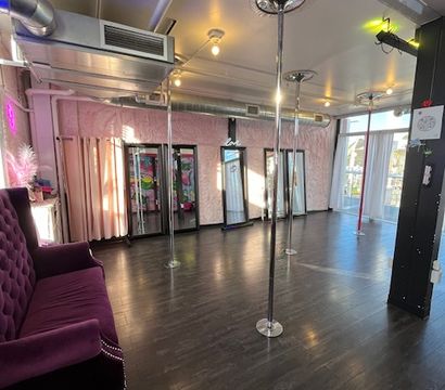 Bright pole fitness studio interior with multiple chrome dance poles, full-length mirrors, dark wood flooring, tufted purple velvet sofa, pink textured walls, exposed ductwork, and floor-to-ceiling windows with natural light.