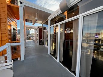 Sunlit waterfront boardwalk-style walkway with glass-front shops, orange container-style facades, metal railing and harbor buildings across the water under a blue sky.