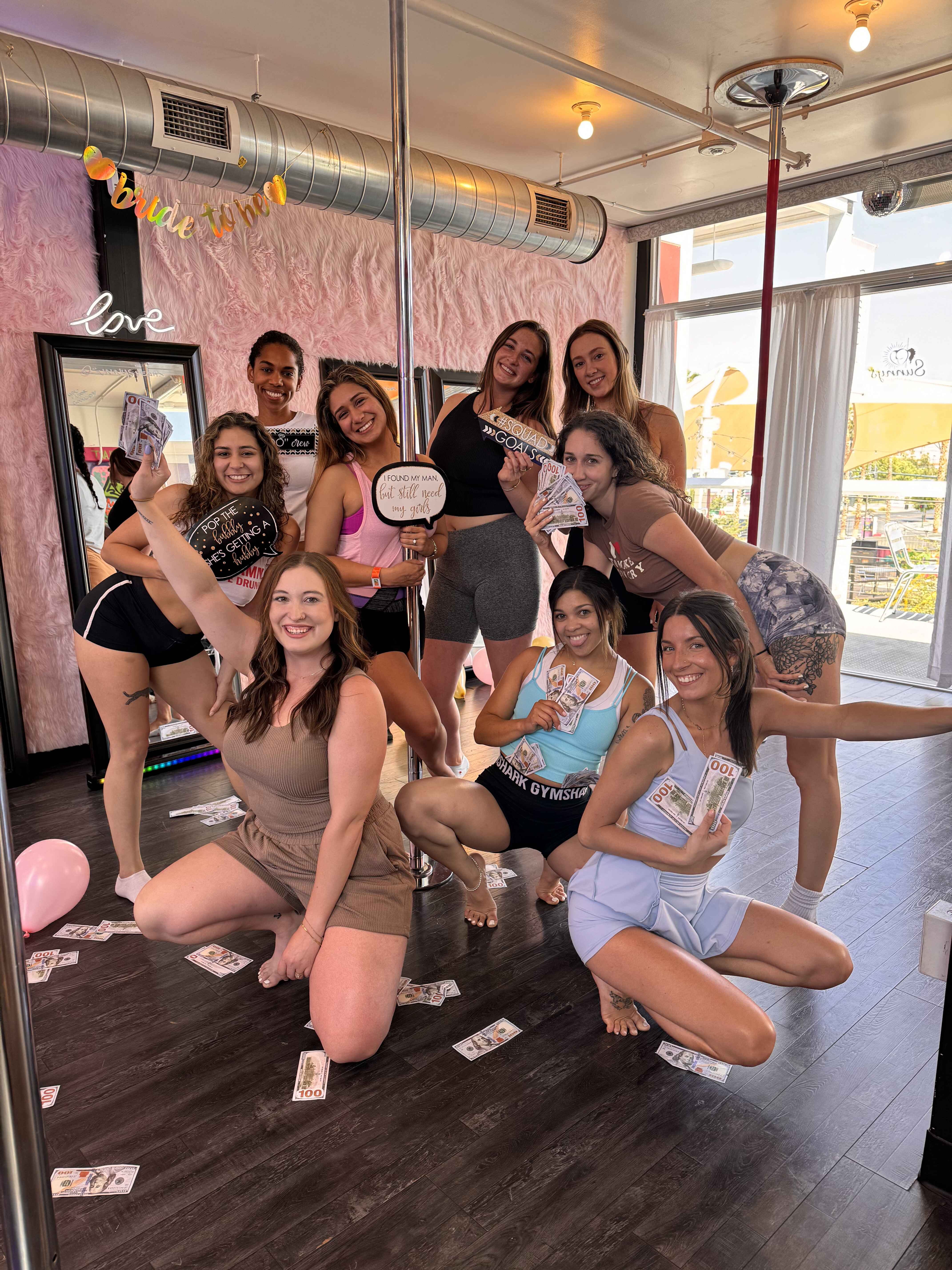 Group of smiling women at a bachelorette pole-dance party in a pink-decor studio, posing with prop $100 bills around a chrome pole, faux-fur wall, mirrors and hardwood floor