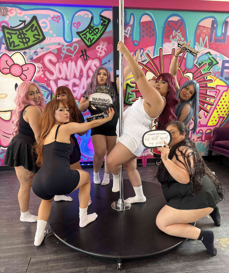 Bachelorette party of seven women posing around a pole in a colorful Vegas-themed pole dance studio with bright graffiti mural, playful photo props and signs.