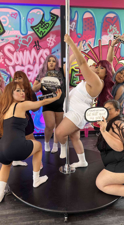 Bachelorette party of seven women posing around a pole in a colorful Vegas-themed pole dance studio with bright graffiti mural, playful photo props and signs.