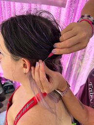 Close-up of a stylist threading purple metallic hair tinsel into a client’s dark hair near the ear, with glitter nails, beaded bracelets and a pink sequin backdrop.