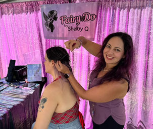 Smiling stylist fitting a delicate hair chain into a seated customer's long ombré hair at a pink sequin vendor booth, with displayed hair extensions and styling tools on a table.