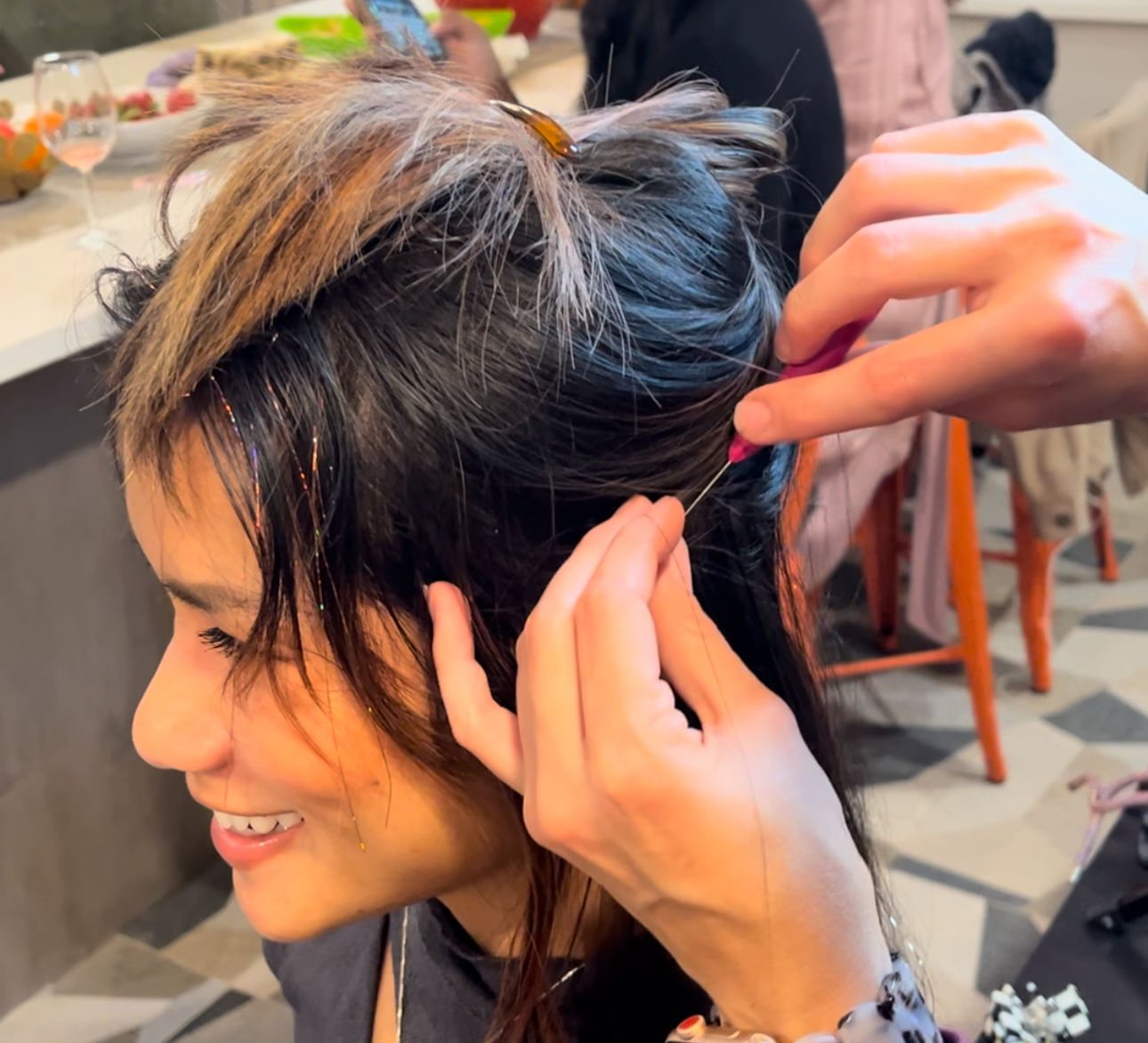 Smiling woman seated indoors as another person threads sparkly hair tinsel into her dark hair with a small tool in a casual salon-style setting.