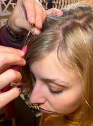 Close-up of blonde hair being decorated with gold hair-tinsel — hands use a pink loop hook to attach thin shimmering strands near the scalp parting.
