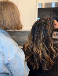 Two people seen from behind — one with a chin-length blonde bob in a denim jacket styling another’s long wavy dark hair with caramel highlights and shimmering tinsel in a home kitchen setting.