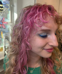 Close-up indoor portrait of a smiling person with pink curly hair, iridescent tinsel highlights, and blue glitter winged eyeshadow by a stained-glass window.