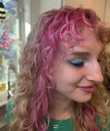 Close-up indoor portrait of a smiling person with pink curly hair, iridescent tinsel highlights, and blue glitter winged eyeshadow by a stained-glass window.