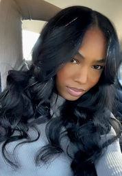 Glam car selfie of a woman with long glossy wavy black hair, soft makeup, and a ribbed gray top, sunlight highlighting her face.