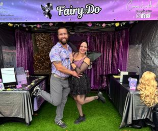 Smiling couple pose with one leg raised in front of a purple fairy-themed vendor booth at an outdoor festival; woman in a black fairy costume with wings and tutu, man in a patterned shirt, wig display and sparkly curtains behind them.