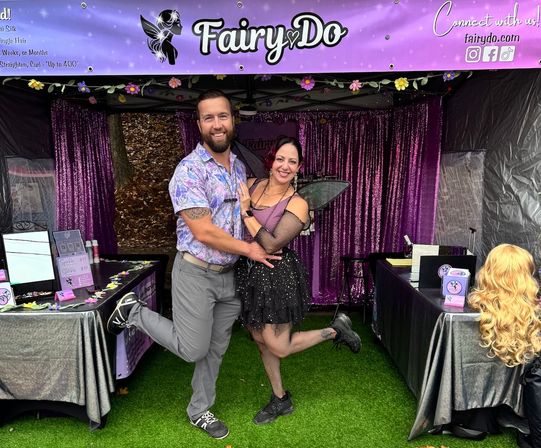 Smiling couple pose with one leg raised in front of a purple fairy-themed vendor booth at an outdoor festival; woman in a black fairy costume with wings and tutu, man in a patterned shirt, wig display and sparkly curtains behind them.