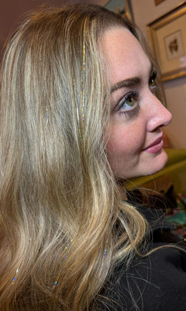 Close-up side profile of a person with long blonde hair and green eyes, delicate iridescent hair-tinsel strands woven through wavy locks against a cozy indoor background.