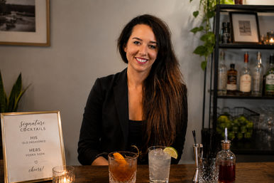 Smiling bartender behind a wooden bar with two cocktails (Old Fashioned and vodka soda), glassware, a signature-cocktails sign, and liquor bottles on shelving in a cozy indoor bar.