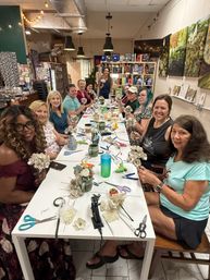 Craft workshop: a group of women seated around a long table in an indoor studio, smiling as they make paper flowers with scissors, glue guns and vases, with bookshelves and artwork in the background.
