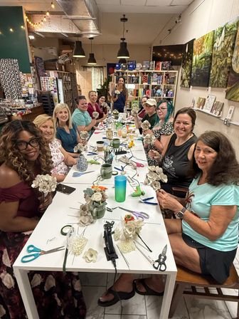 Craft workshop: a group of women seated around a long table in an indoor studio, smiling as they make paper flowers with scissors, glue guns and vases, with bookshelves and artwork in the background.