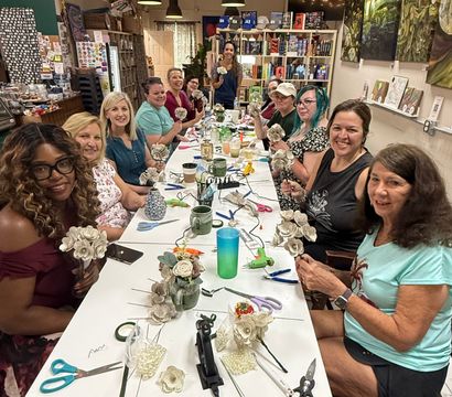 Craft workshop: a group of women seated around a long table in an indoor studio, smiling as they make paper flowers with scissors, glue guns and vases, with bookshelves and artwork in the background.