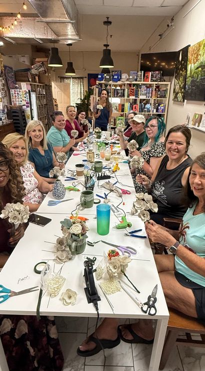 Craft workshop: a group of women seated around a long table in an indoor studio, smiling as they make paper flowers with scissors, glue guns and vases, with bookshelves and artwork in the background.
