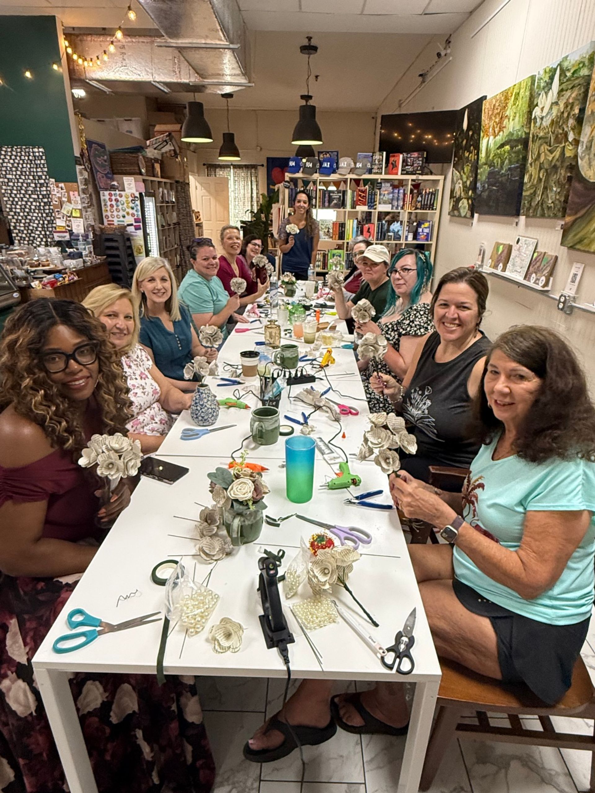 Craft workshop: a group of women seated around a long table in an indoor studio, smiling as they make paper flowers with scissors, glue guns and vases, with bookshelves and artwork in the background.