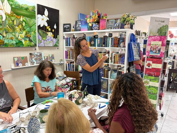 Instructor demonstrating paper-flower craft to a small group at a cozy independent bookstore workshop, surrounded by bookshelves, a greeting-card rack, colorful floral artwork and craft supplies on the table.