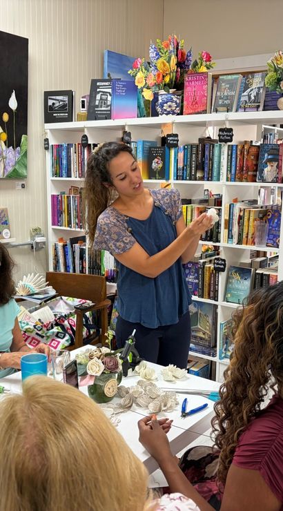 Instructor demonstrating paper-flower craft to a small group at a cozy independent bookstore workshop, surrounded by bookshelves, a greeting-card rack, colorful floral artwork and craft supplies on the table.