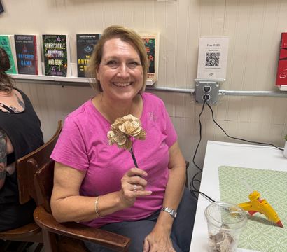 Smiling woman in a pink shirt holding a handmade paper flower made from book pages at an indoor craft workshop, with books and paintings on the wall and a glue gun on the table.