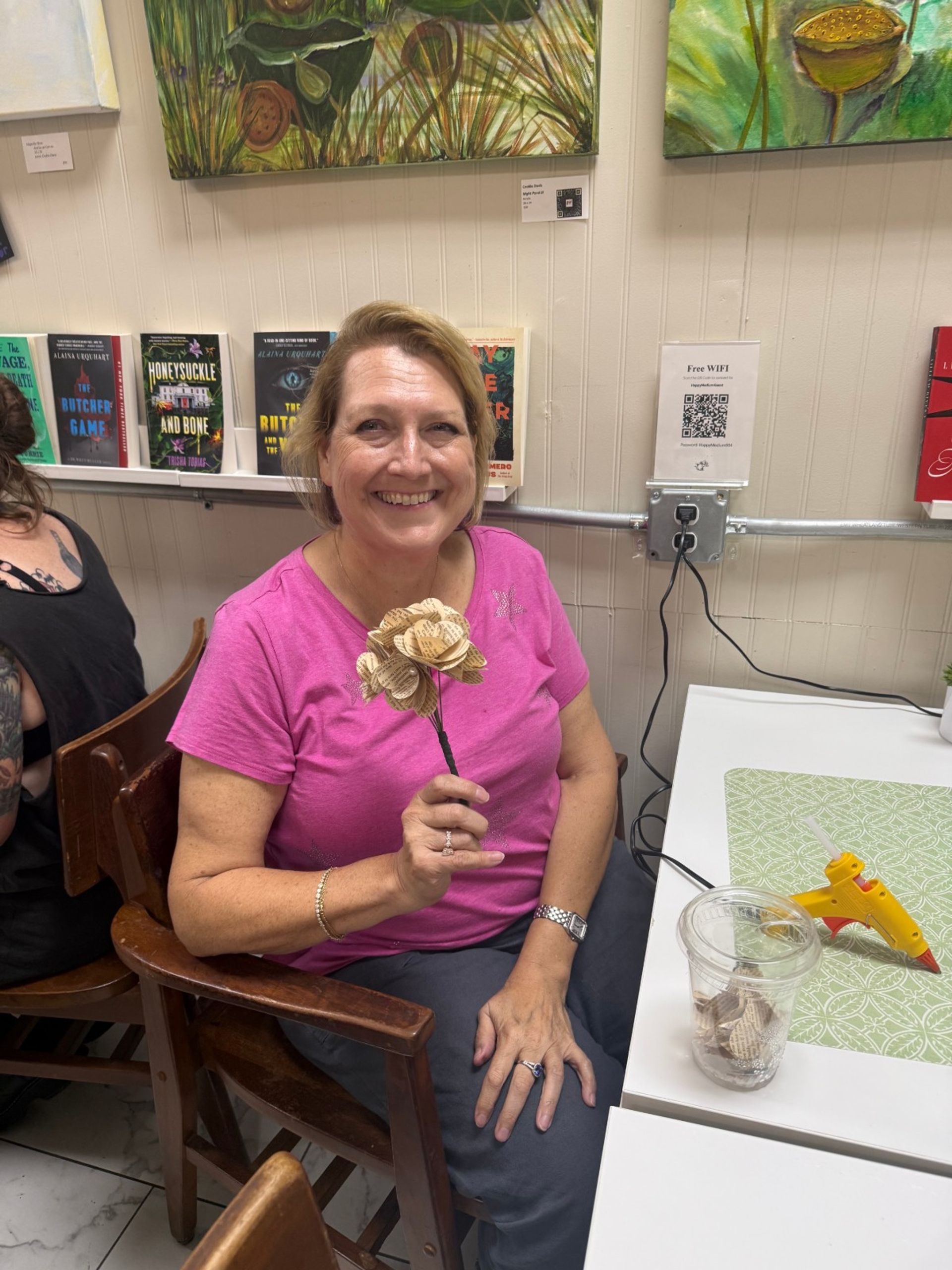 Smiling woman in a pink shirt holding a handmade paper flower made from book pages at an indoor craft workshop, with books and paintings on the wall and a glue gun on the table.