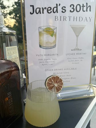 Outdoor cocktail setup with a printed birthday drinks menu showing Yuzu Margarita and Lychee Martini, a pale yellow yuzu margarita in a ribbed glass garnished with a dried lime wheel, and liquor bottles with a sunset background.