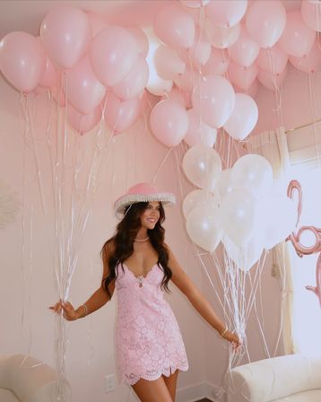 Smiling woman in a pink lace dress and fringed hat holding clusters of pale pink and white helium balloons in a pastel pink indoor party scene