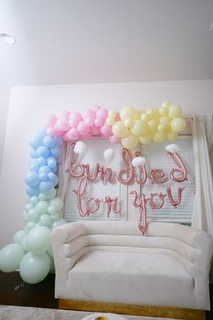 Cheery pastel balloon arch (pink, yellow, blue, mint) with rose-gold letter balloons spelling bundled for you and cotton cloud accents above a cream velvet loveseat — indoor baby shower backdrop