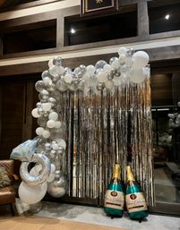 Indoor silver-and-white balloon arch with clear and metallic balloons, a holographic ring balloon and two oversized champagne bottle balloons against a silver tinsel fringe photo backdrop in a wood-paneled lounge