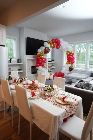 Open-plan modern dining and living room decorated for a strawberry-themed party with a white-linen table, red-and-white runner, gold-rimmed plates, floral centerpieces, champagne, and a large circular red, white and gold balloon arch with strawberry balloons by the windows and TV.