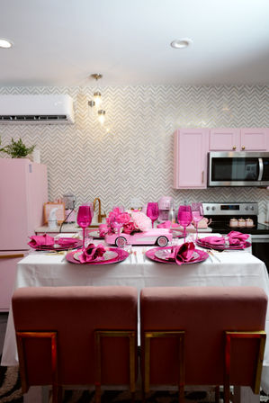 Vibrant pink-themed modern kitchen with chevron tile backsplash, pastel pink cabinets and fridge, a white table set for four with fuchsia plates, napkins and wine glasses, pink floral centerpiece with retro toy-car accent and gold-accent dining chairs.