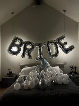 Playful bridal surprise in a modern bedroom: large silver foil balloons spelling "BRIDE" suspended above a bed scattered with white and metallic balloons under soft pendant lighting.