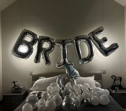 Playful bridal surprise in a modern bedroom: large silver foil balloons spelling "BRIDE" suspended above a bed scattered with white and metallic balloons under soft pendant lighting.