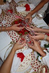 Overhead view of hands toasting with clear shot glasses at a strawberry-themed summer table featuring a basket of fresh strawberries, strawberry-patterned tablecloth, floral vintage china, gold flatware and red-trimmed napkins