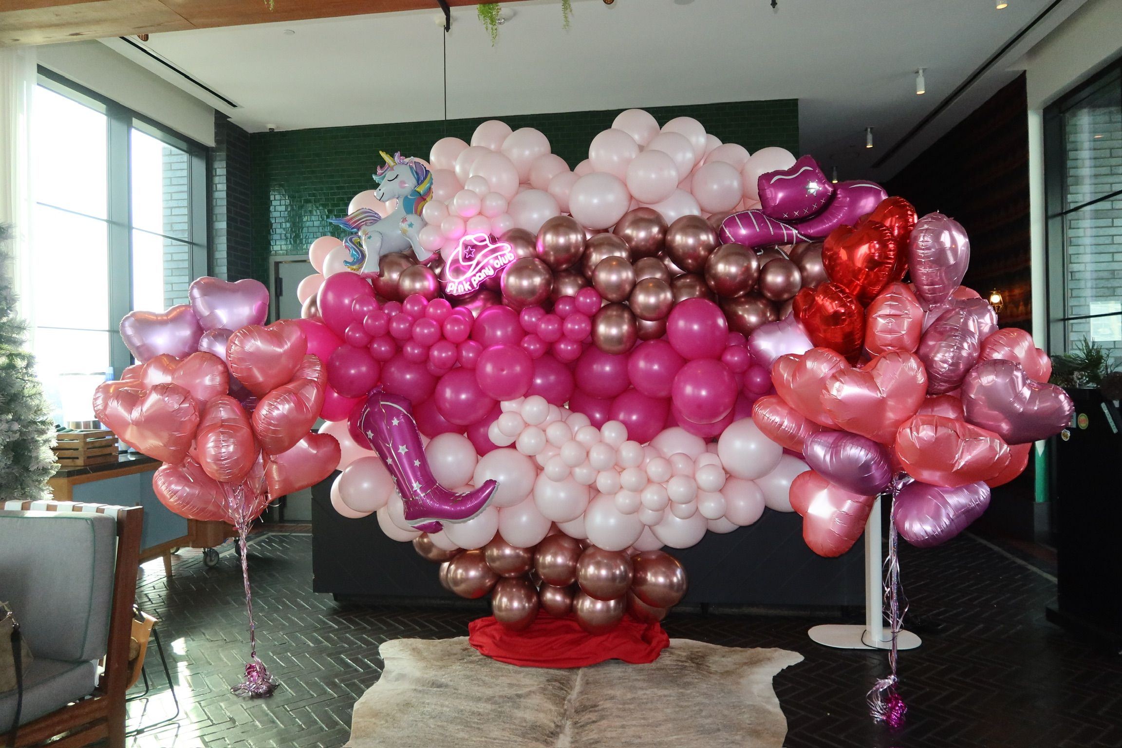 Bright pink and rose-gold balloon backdrop shaped like giant lips with heart-shaped foil balloon bouquets and unicorn and boot foil accents — festive party photo backdrop in a modern indoor event space.